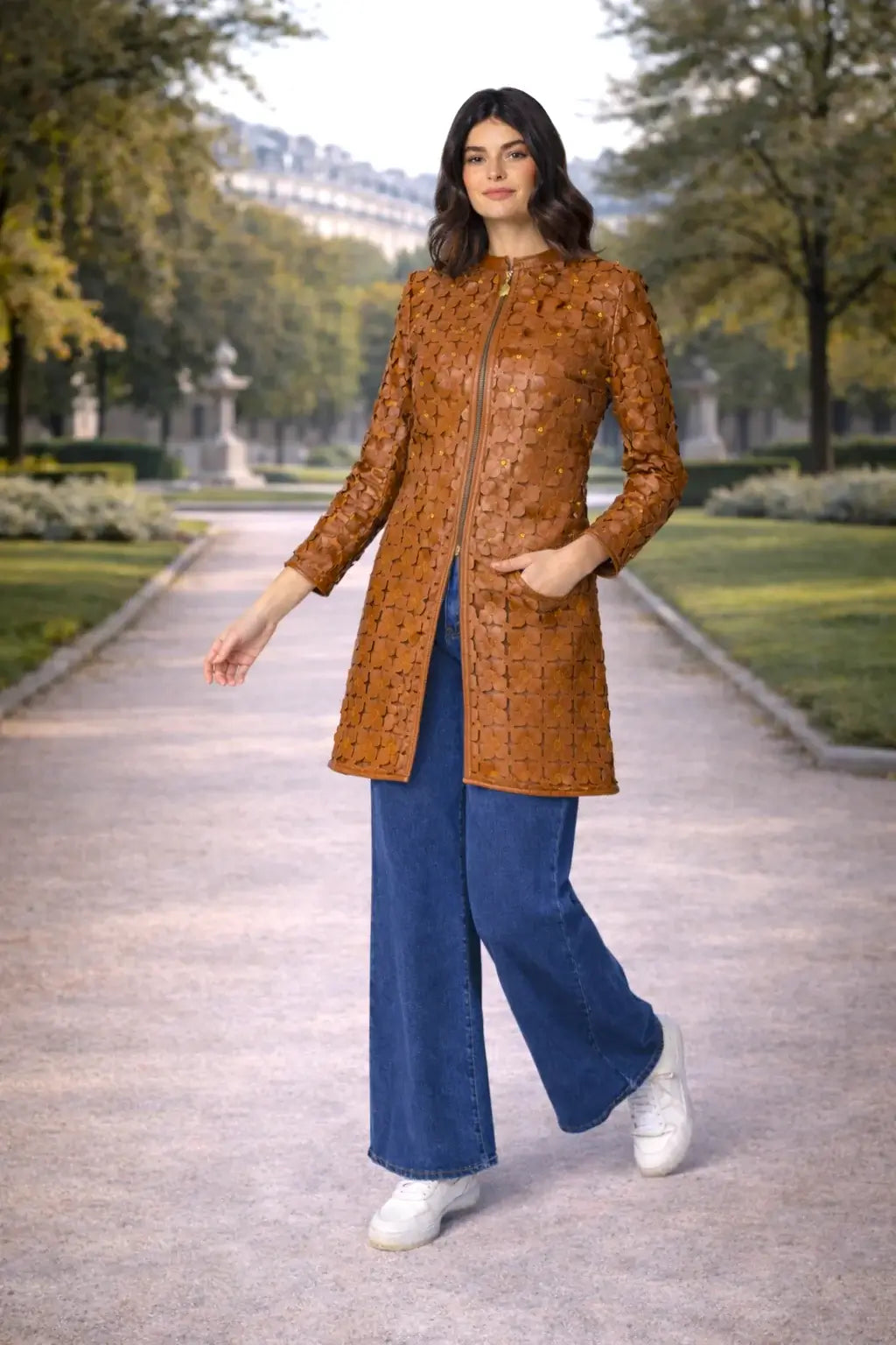 Modella che indossa la giacca LUNA in pelle marrone, abbinata a pantaloni blu e sneakers, in un parco elegante.